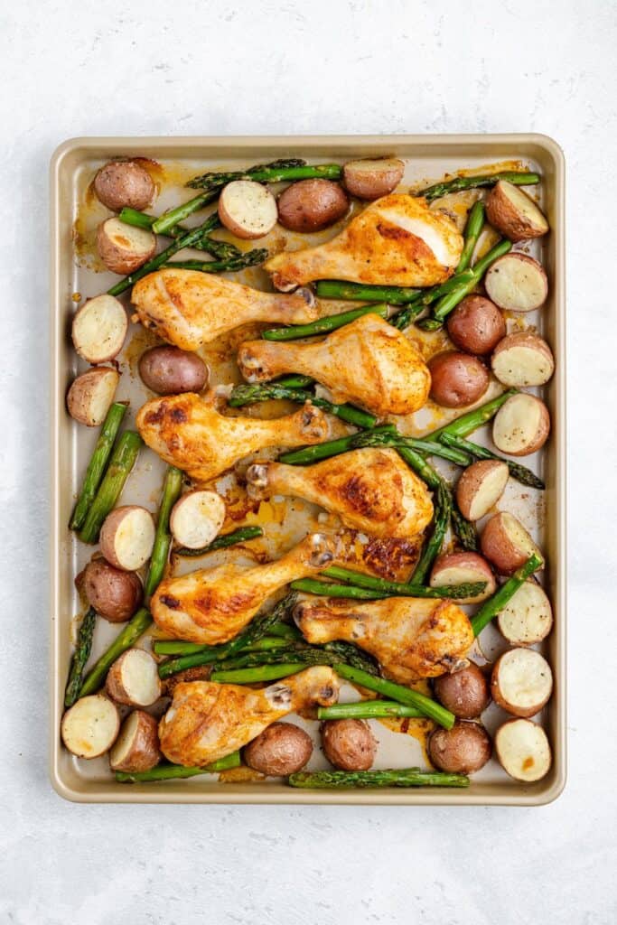 Chicken drumsticks, red potatoes, and asparagus spears on a sheet pan atop a light-colored surface.