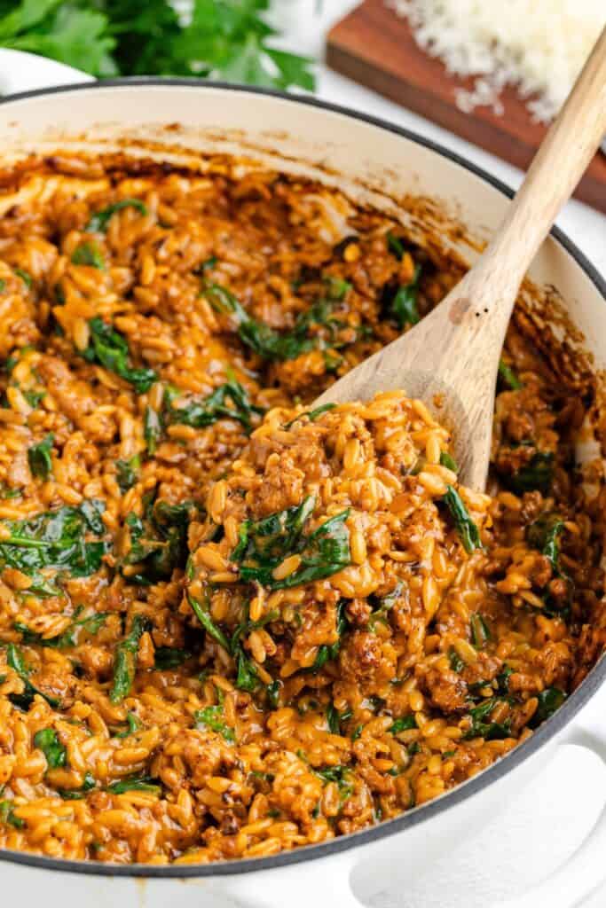 Wooden spoon scooping creamy orzo with sausage and spinach out of a pot.