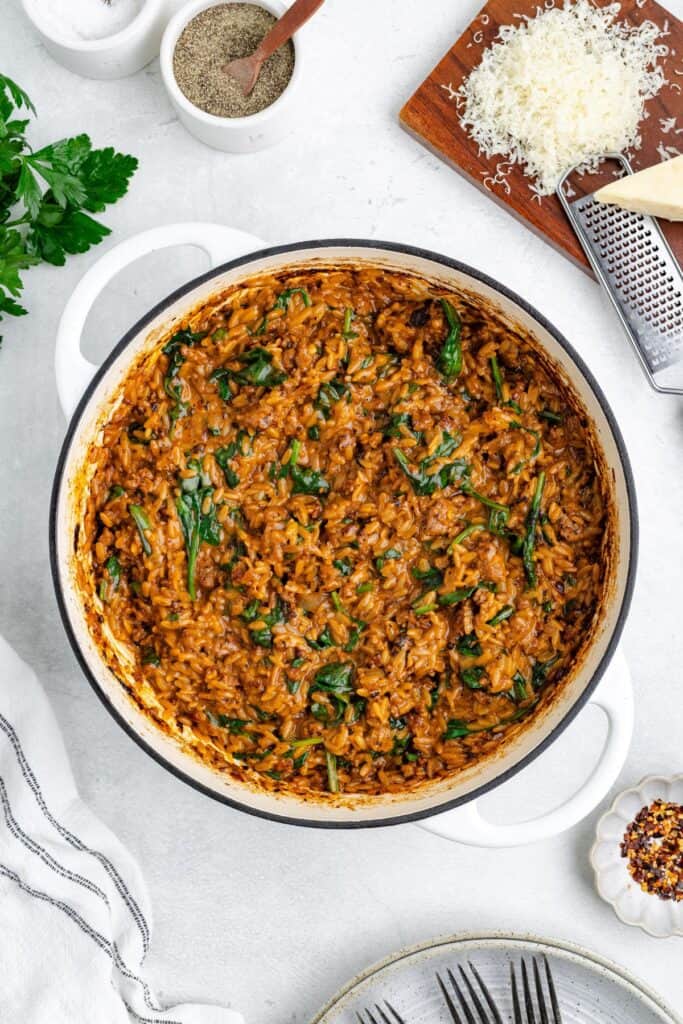 A pot of creamy Italian sausage and orzo, surrounded by cheese, pepper grinder, parsley, and a striped towel.