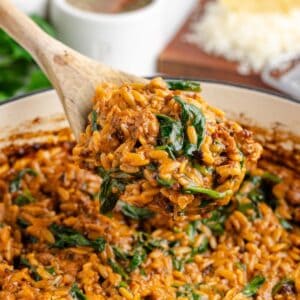 A wooden spoon lifts creamy sausage and orzo with spinach from a pot.