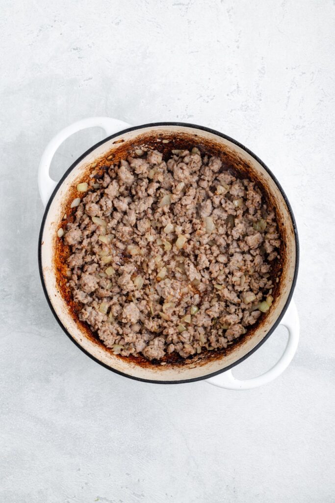 White enameled pot with browned ground meat and chopped onions on a light gray textured background.