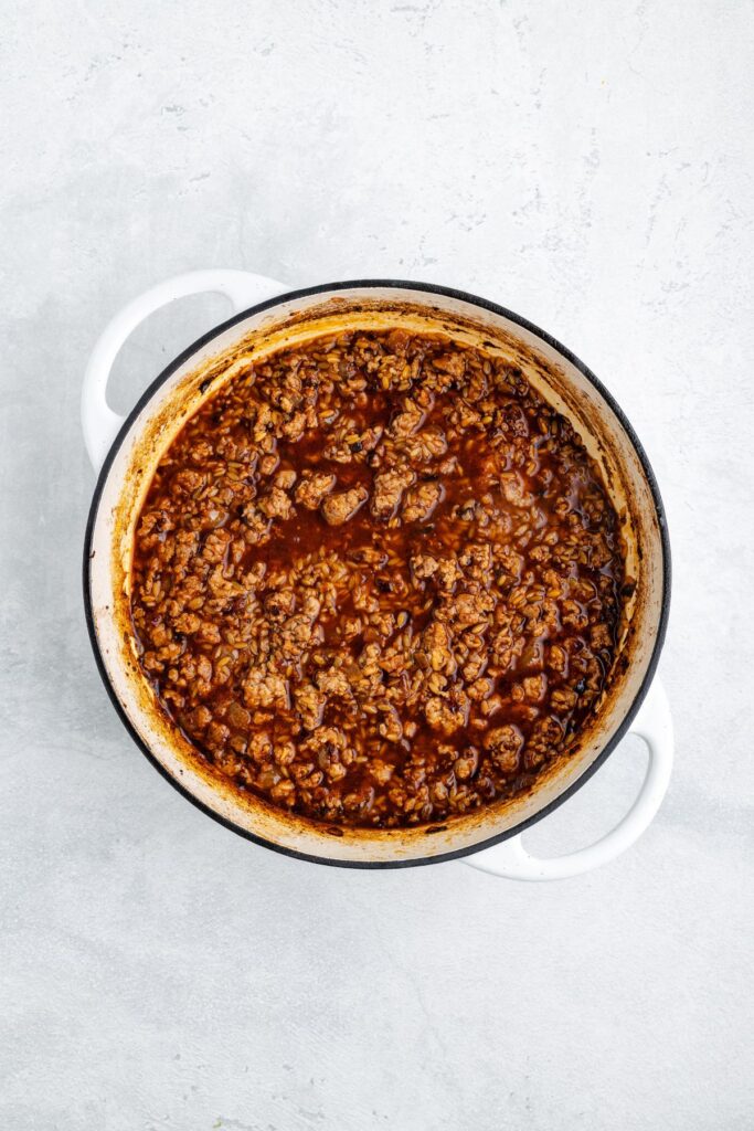 A white pot holding cooked ground beef in reddish-brown sauce, placed on a light gray surface.