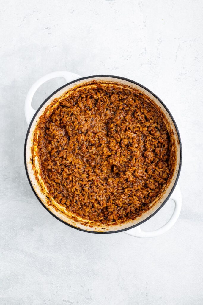 White pot containing cooked rice and dark reddish-brown sauce, placed on a light gray surface.