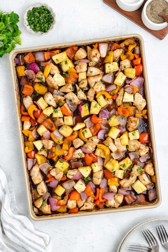 Sheet pan of cooked diced chicken, pineapple, red onions, and bell peppers with herbs on a white surface.