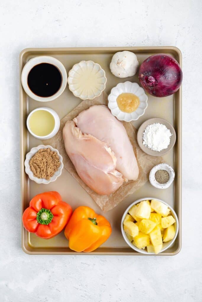 Baking tray with raw chicken breasts surrounded by pineapple, bell peppers, red onion, spices, and sauces in dishes.