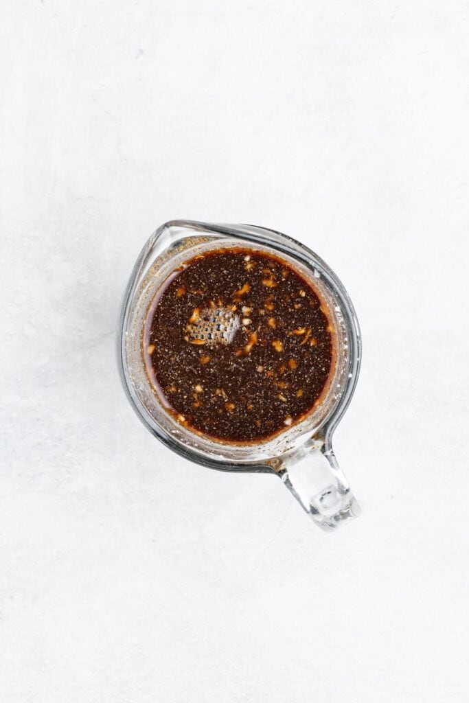 Glass measuring cup with dark brown marinade and visible spices, top-down view on a light background.