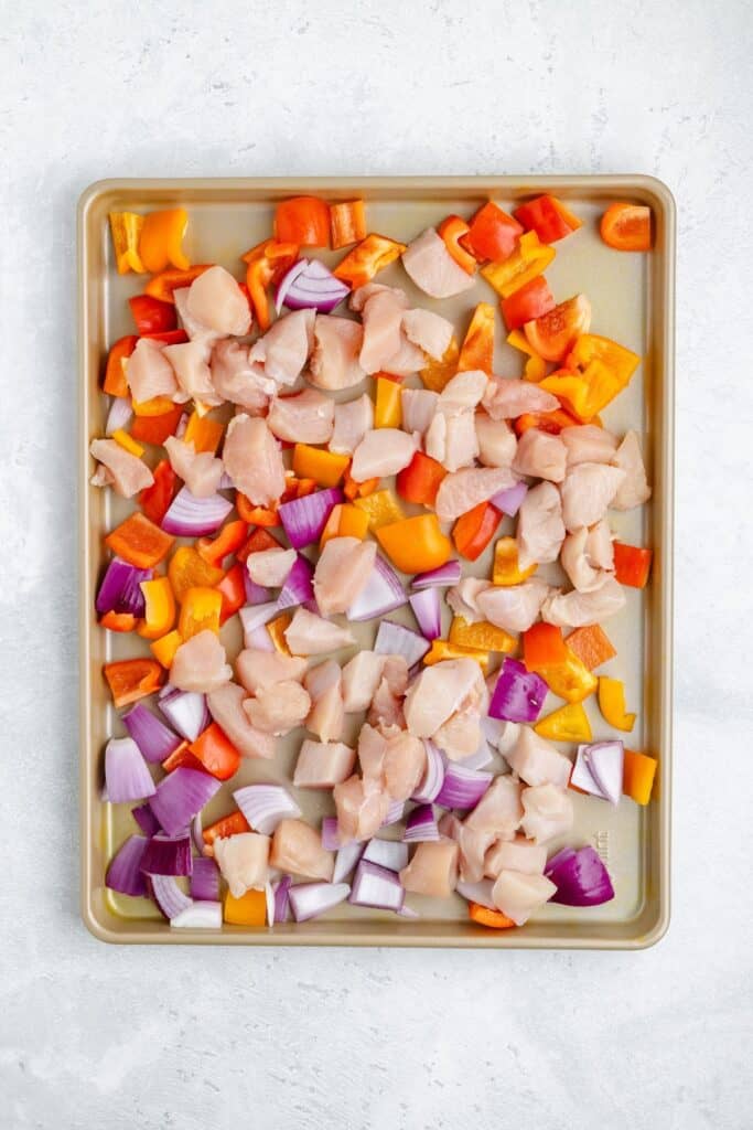 Evenly arranged raw chicken, red onion, and orange and red bell pepper pieces on a baking sheet.
