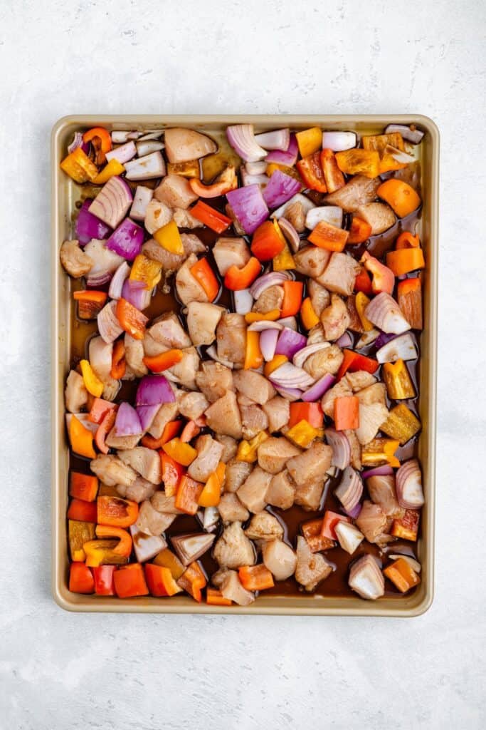Raw chopped chicken, red onions, and orange and yellow bell peppers on a baking sheet ready for roasting.