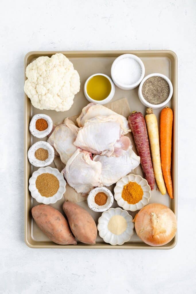 Raw chicken thighs, assorted vegetables, and spices arranged on a baking sheet with olive oil, salt, and pepper.