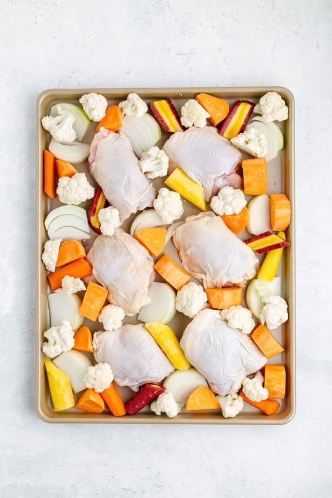Baking sheet with raw chicken thighs, carrots, sweet potatoes, cauliflower, and onion on a light surface.