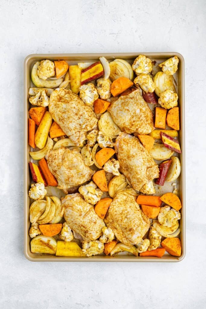 Baking sheet with five seasoned chicken thighs and assorted chopped vegetables, ready to roast.