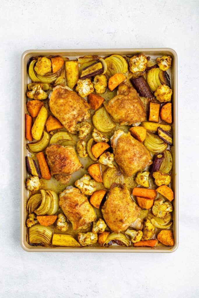 Sheet pan with four seasoned chicken thighs and assorted roasted vegetables on a light background.