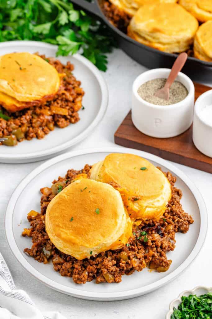 Two plates of ground beef sloppy Joe casserole with biscuits, plus a skillet of the dish and salt and pepper shakers nearby.