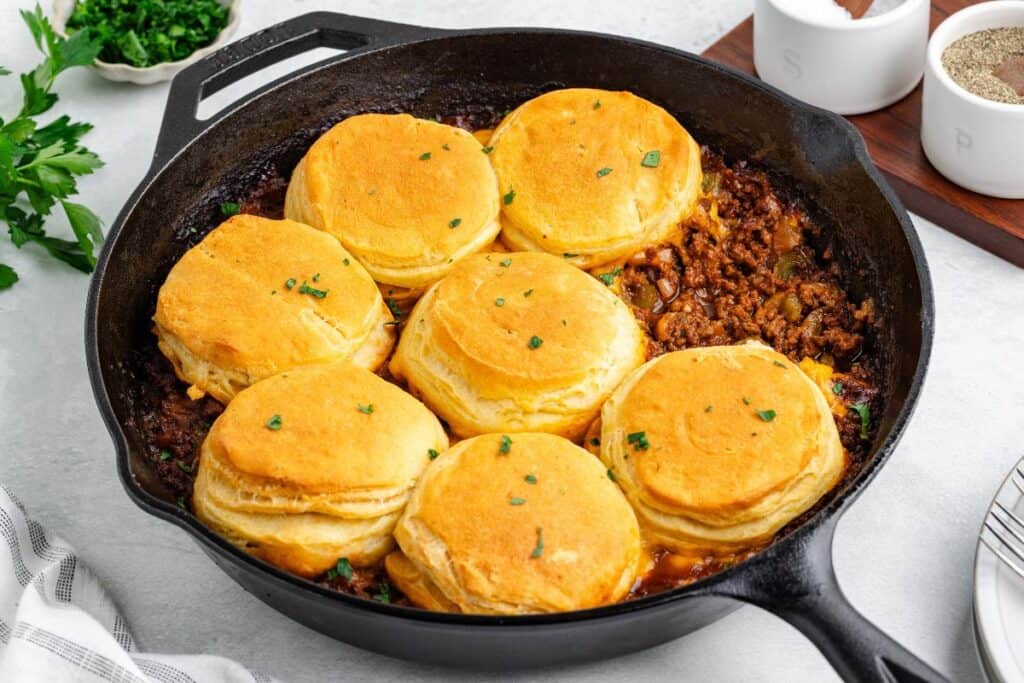 Cast iron skillet of biscuit sloppy Joe casserole with seven golden-brown biscuits arranged on top.