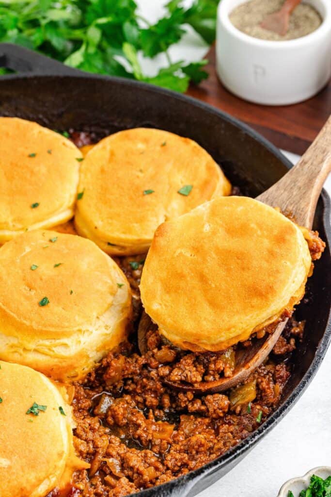 Cast iron skillet of sloppy Joe biscuit casserole; wooden spoon lifts a biscuit and filling.