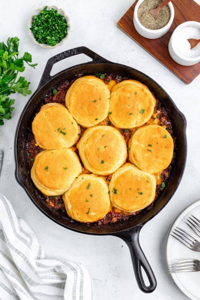Cast iron skillet with baked biscuits atop meat and vegetable filling, garnished with parsley. Plates nearby.