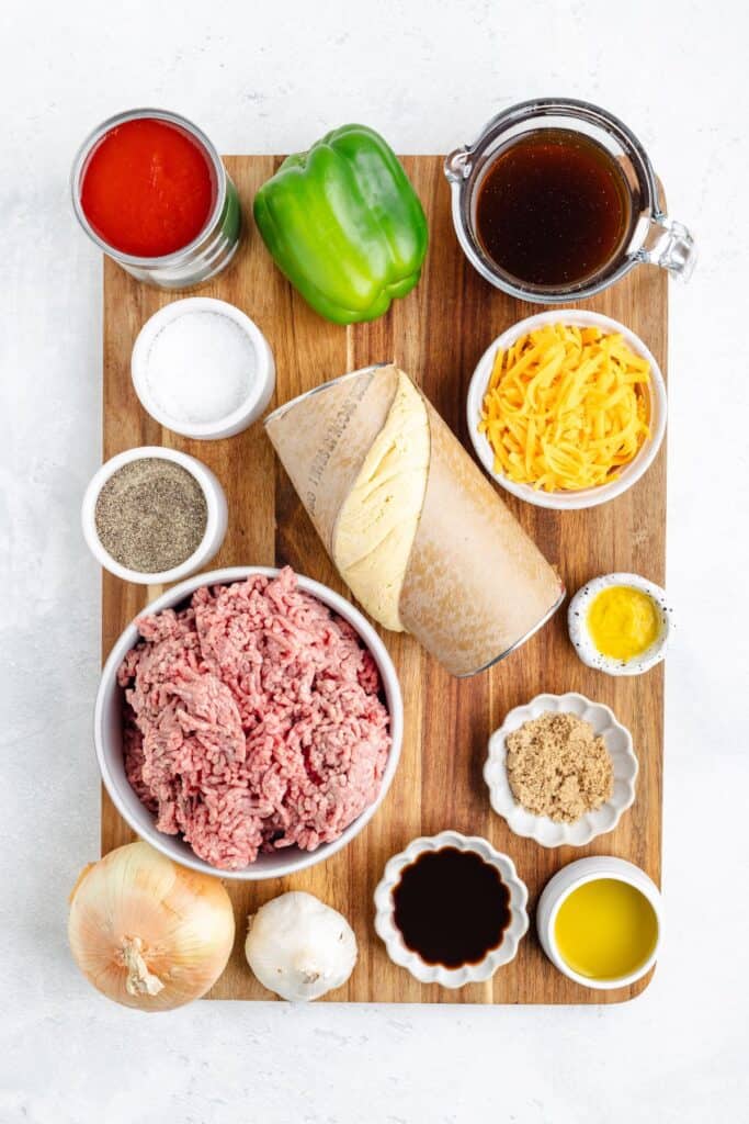 Ingredients on a wooden board: ground beef, hoagie roll, cheese, bell pepper, onion, garlic, sauces, seasonings.