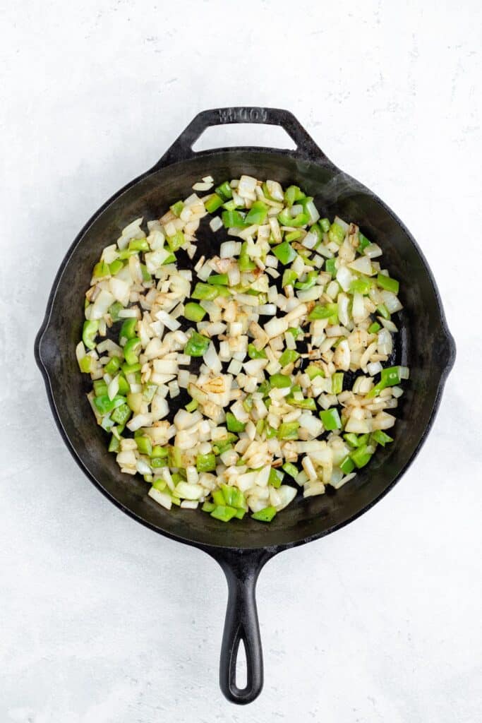 Diced onions and green bell peppers sauté in a black cast iron skillet on a light countertop.
