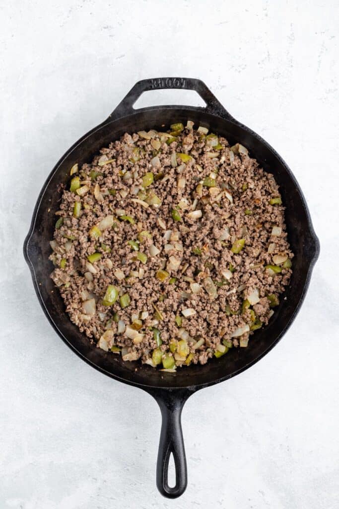 Cast iron skillet with cooked ground beef, chopped onions, and green bell peppers on a light background.