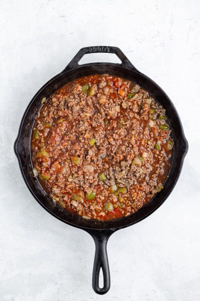 Cast iron skillet with cooked ground beef, green bell peppers, onions, and tomato sauce on a light surface.