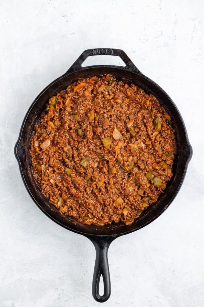 Cast iron skillet with ground beef, green bell peppers, onions, and tomato sauce on a light surface.