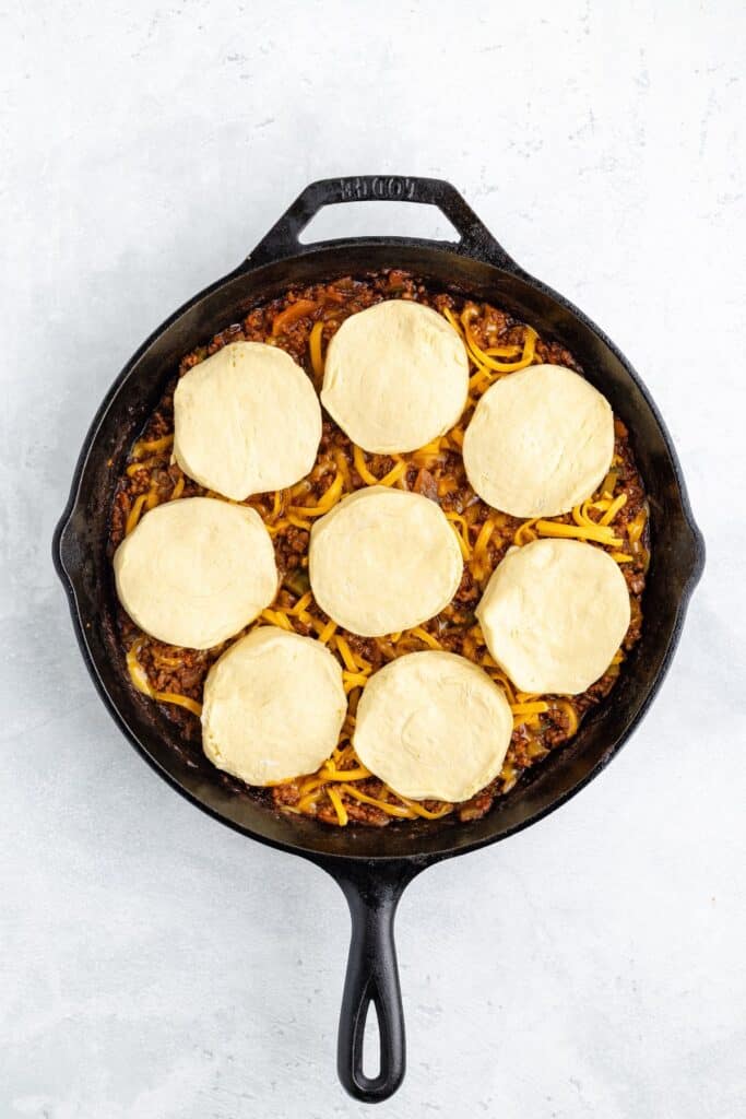 Cast iron skillet with ground meat, vegetables, shredded cheese, and eight unbaked biscuit rounds on top.