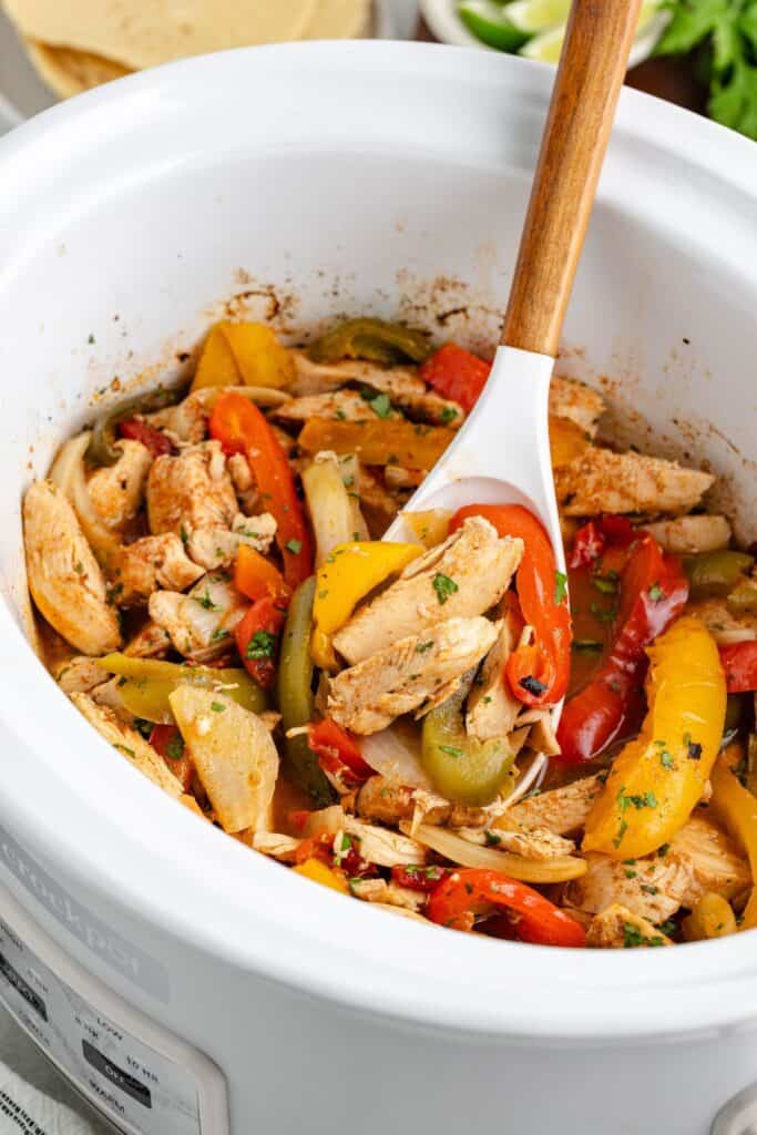 Crockpot chicken fajitas, colorful bell peppers, and onions cooking in a white slow cooker with a wooden spoon.