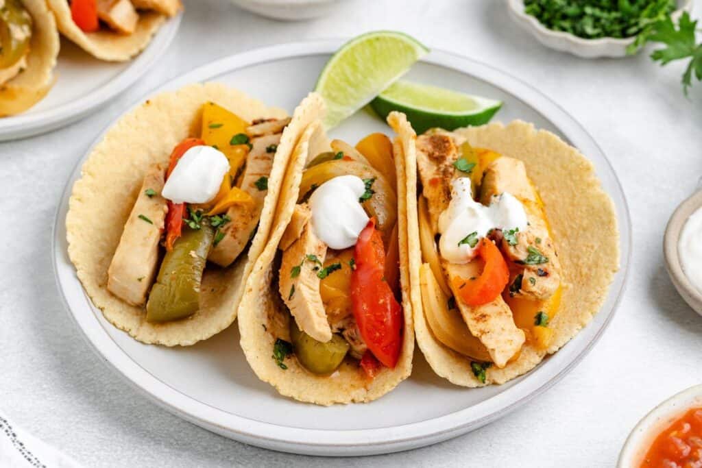 Slow Cooker Chicken fajitas in tortillas with bell peppers, onions, and sour cream on corn tortillas, with lime wedges on a white plate.