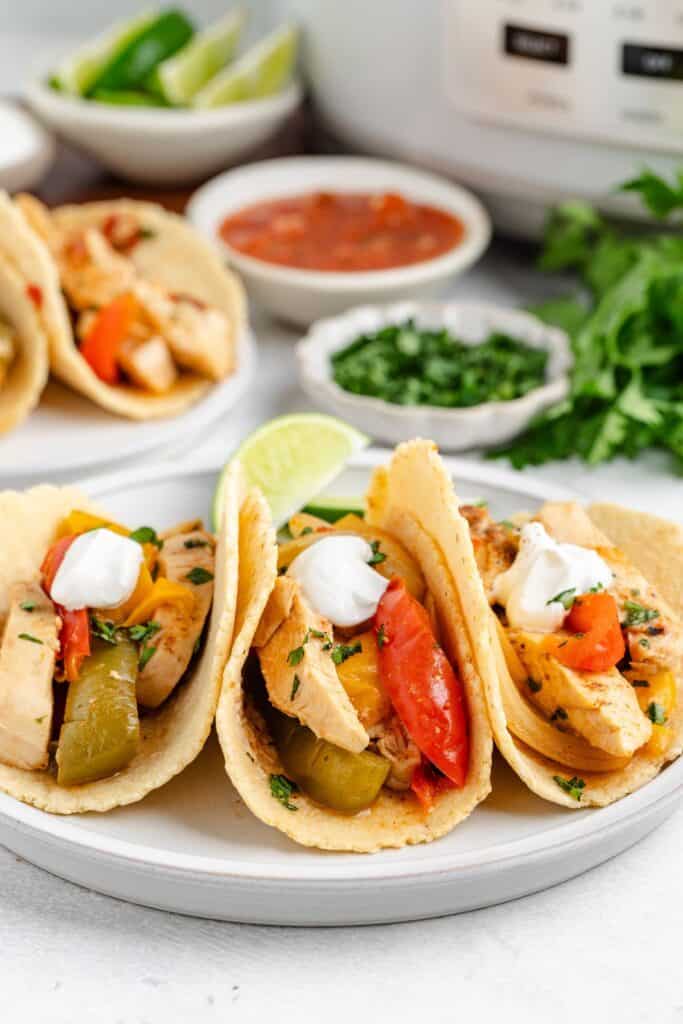 Three tortillas with crockpot chicken fajitas with bell peppers, onions, and sour cream on a plate with a lime wedge.