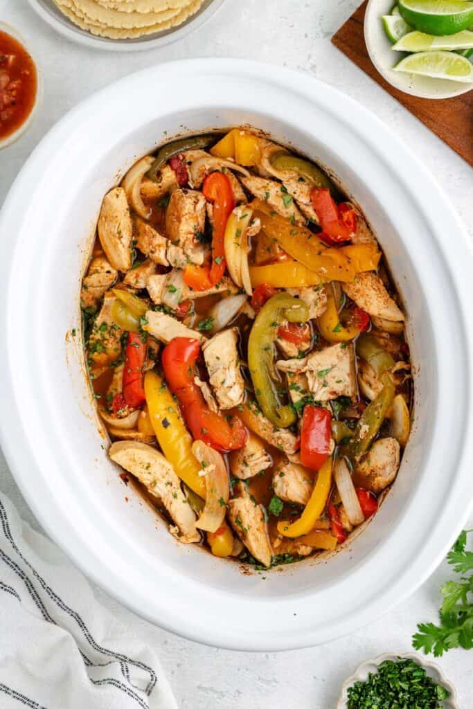 Chicken Fajitas in the Slow Cooker with tortillas, lime wedges, salsa, and herbs nearby.