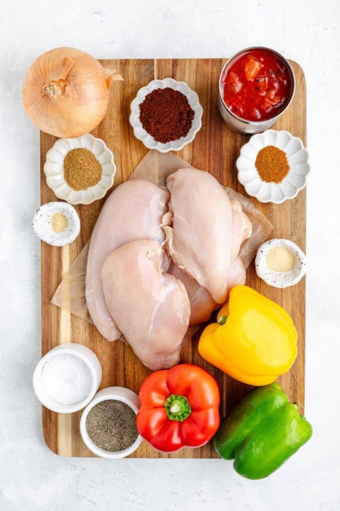 Raw chicken, bell peppers, onion, canned tomatoes, and spices arranged on a wooden cutting board for cooking prep.