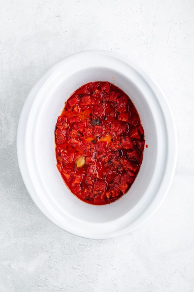White slow cooker containing chunky tomato sauce, placed on a light gray surface.