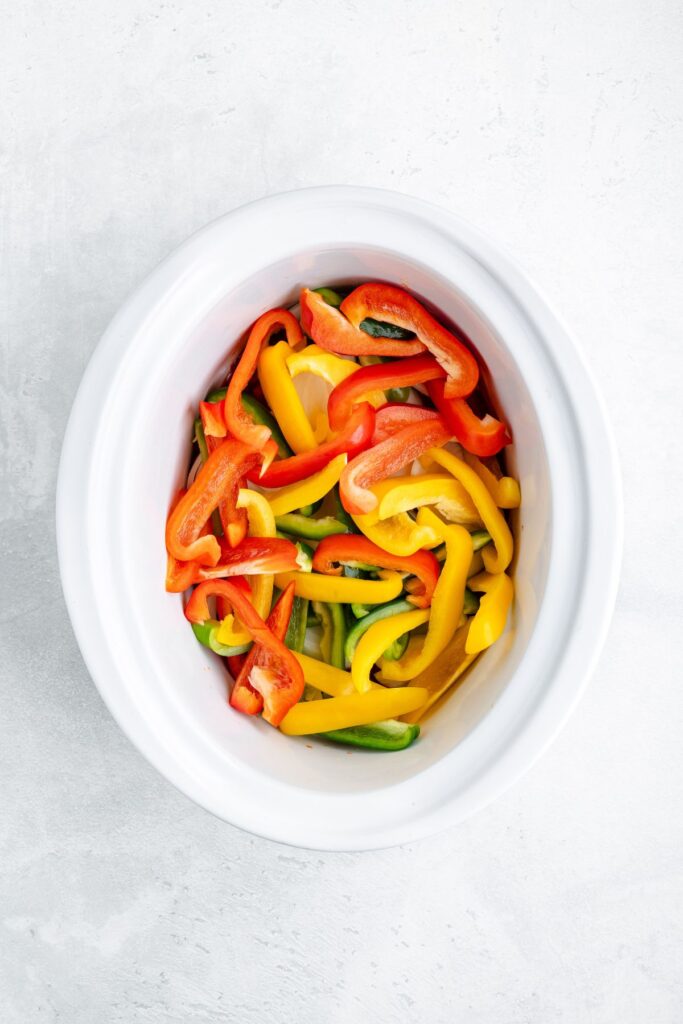 Sliced red, yellow, and green bell peppers in a white slow cooker on a light gray surface.