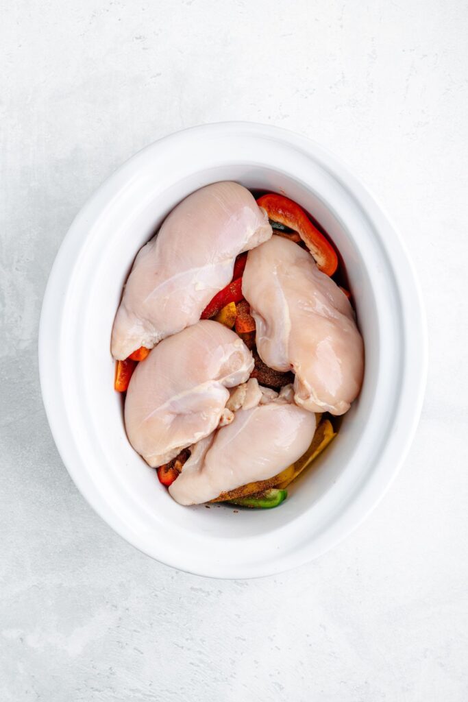 Four raw chicken breasts on sliced bell peppers and onions in a white slow cooker on a light surface.