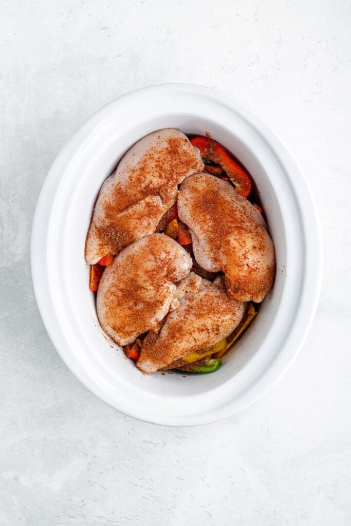 Seasoned raw chicken breasts on sliced vegetables in a white oval slow cooker, set on a light surface.