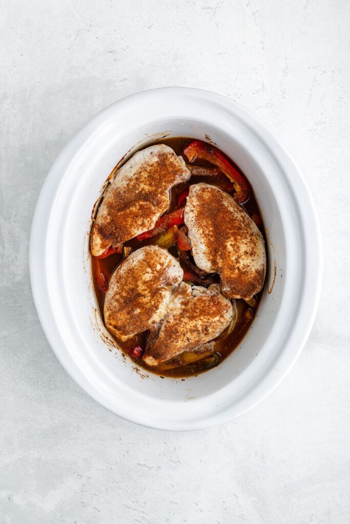 Seasoned chicken breasts and vegetables in a slow cooker on a light countertop.