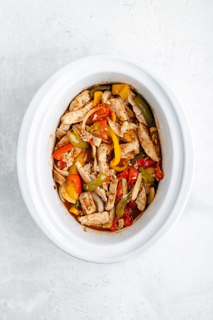White slow cooker with sliced cooked chicken, bell peppers, and onions on a light gray surface.