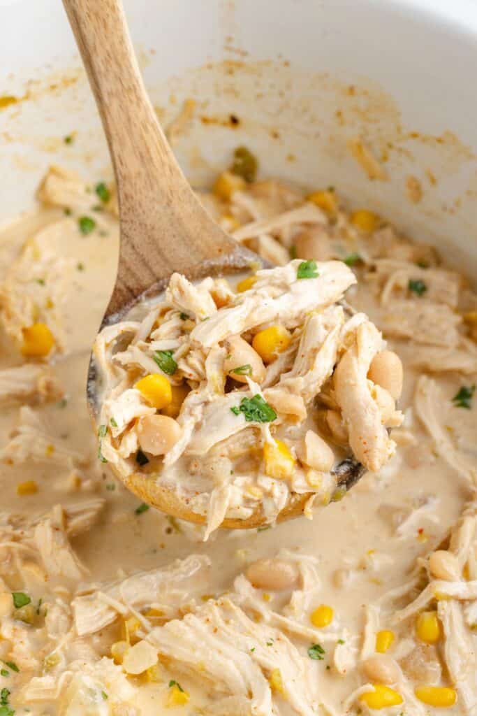 Wooden spoon lifting creamy chicken chili with corn, and white beans with herbs from a slow cooker.