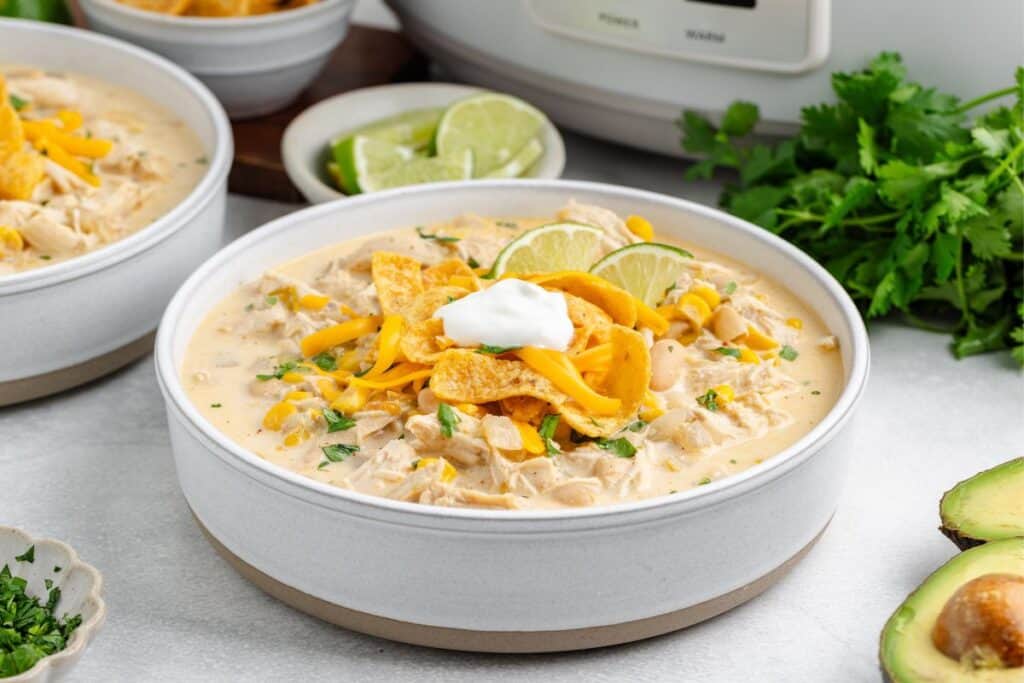 Bowl of crockpot white chicken chili with cheese, tortilla chips, lime, sour cream, herbs, and avocado.