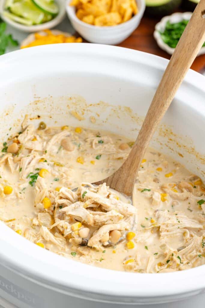 Slow cooker chicken white chili with corn and beans in creamy broth being stirred with a wooden spoon in a crockpot.
