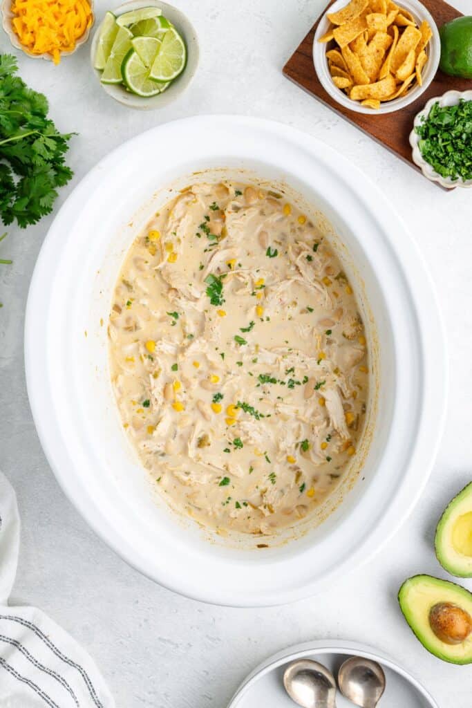 A slow cooker of creamy white chicken chili with cilantro, surrounded by bowls of toppings and avocado halves.