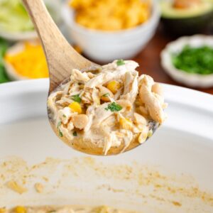 Wooden spoon with creamy crockpot chicken white chili over a slow cooker; blurred toppings in background.