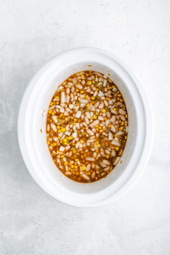 White slow cooker containing chopped onions, minced garlic, and brown marinade on a light textured surface.