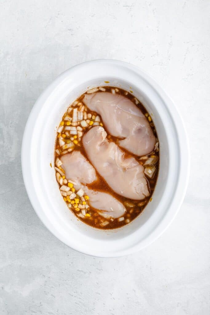 Four raw chicken breasts with diced onions and brown marinade in a white slow cooker on a gray surface.