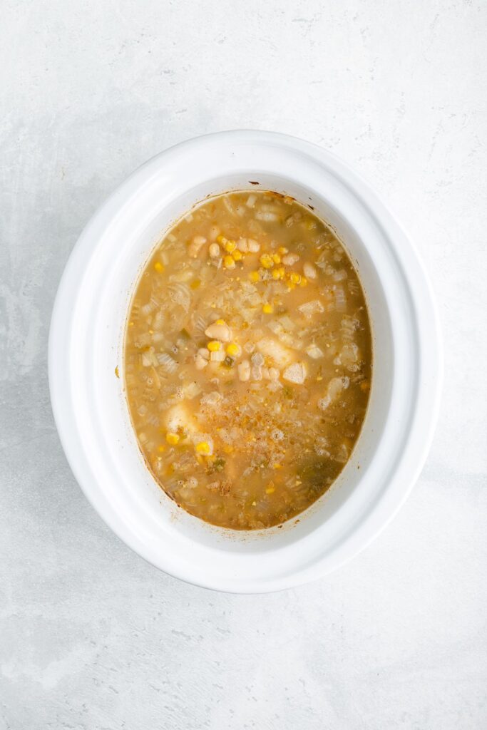 White slow cooker with light brown soup, corn, beans, and vegetables on a white surface.