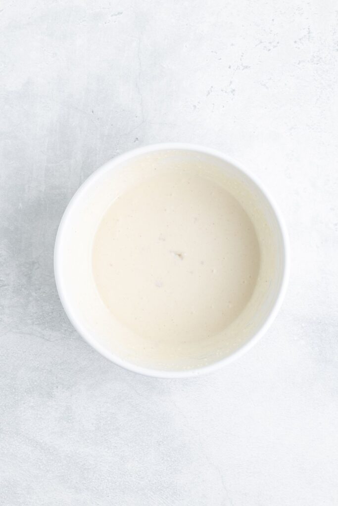 White bowl with creamy, beige sauce or batter on a light gray textured surface.