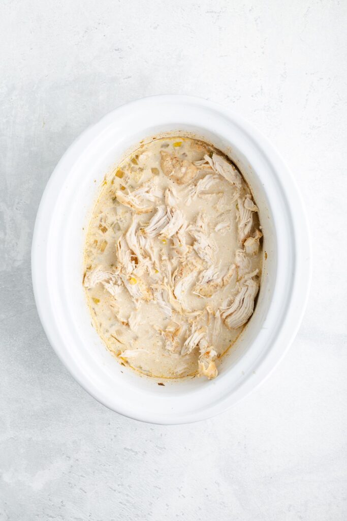 Shredded chicken in creamy sauce inside a white slow cooker, viewed from above on a light background.