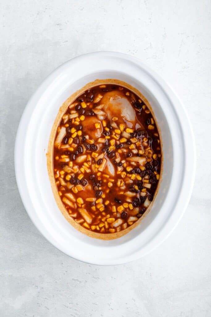 Raw chicken breasts in a white slow cooker topped with black beans, corn, onions, and red sauce.