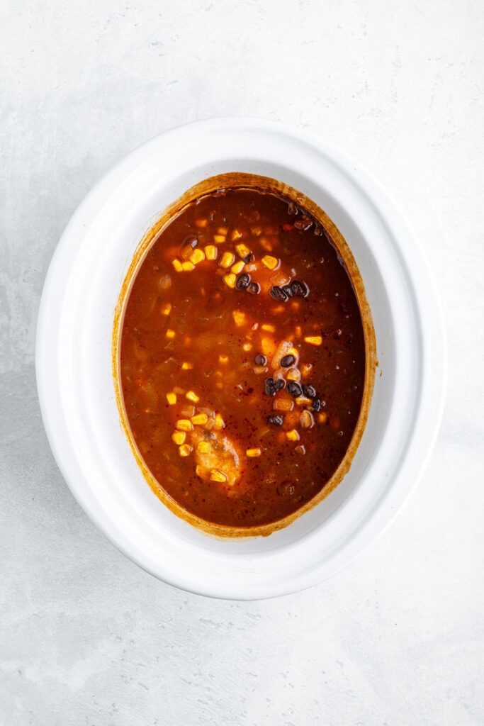 White slow cooker with corn, black beans, and chicken soup in red broth, viewed from above.