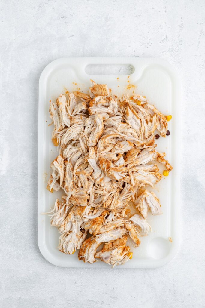 Shredded seasoned chicken on a white cutting board set on a light surface.
